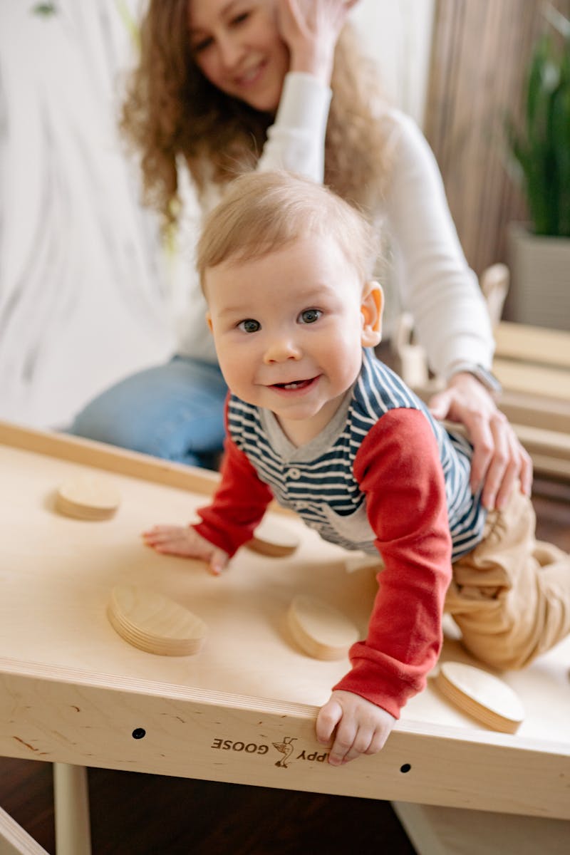 Krabbelgruppe, Baby, Kind, Adorable baby crawling playfully on a wooden toy table with supportive mother nearby, indoors.