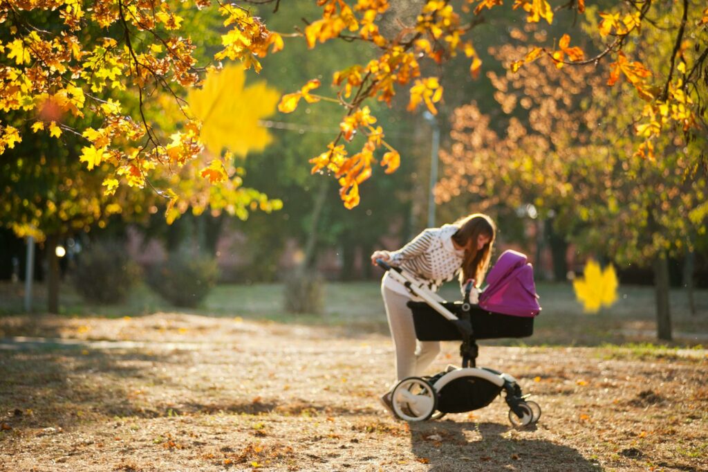 Mother strolling in a sunny autumn park with a baby stroller, surrounded by colorful leaves.